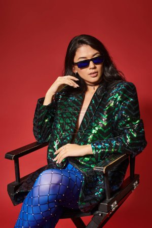 pretty asian woman in sunglasses and green jacket with sequins sitting on chair on red backdrop