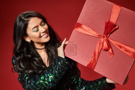 smiling asian woman in jacket with sequins and blue pantyhose holding present on red backdrop