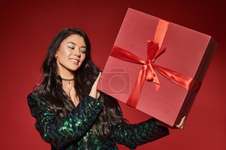 glückliche asiatische Frau in grünem Sakko mit Pailletten hält Geschenk auf rotem Hintergrund, Frohe Weihnachten