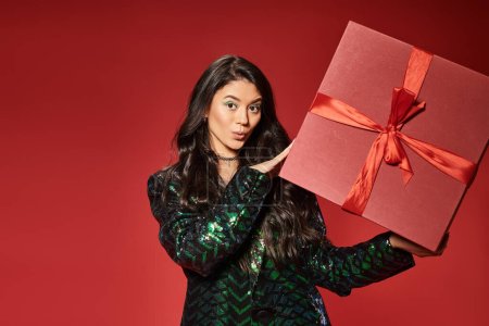surprised asian woman in green jacket with sequins holding gift box on red backdrop, Merry Christmas