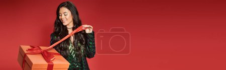 happy asian woman in green jacket with sequins pulling ribbon on gift box, Merry Christmas banner