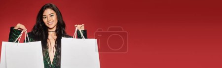 Photo for Black friday banner, joyful asian woman in green jacket with sequins holding shopping bags on red - Royalty Free Image