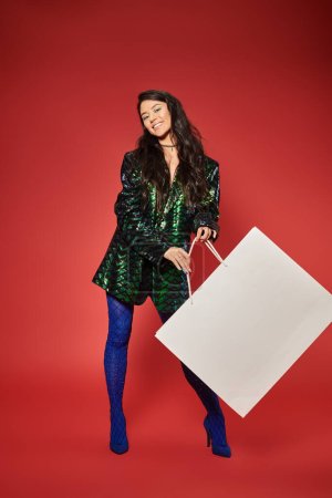 black friday, joyful asian woman in green jacket with sequins holding huge shopping bag on red