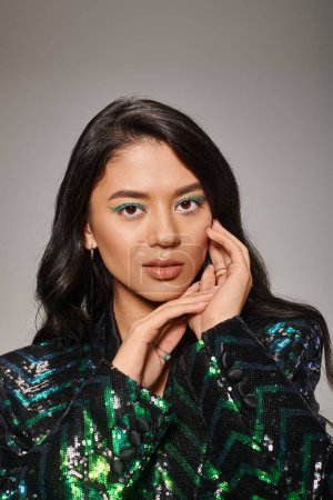 brünette asiatische Frau in grünem Sakko mit Pailletten und fettem Augen-Make-up posiert vor grauem Hintergrund