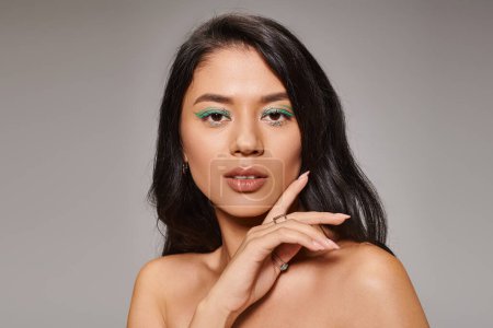 beautiful asian woman with green eyeliner and bare shoulders posing on grey background, bold makeup