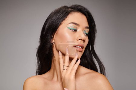 brunette asian woman with green eyeliner and bare shoulders posing on grey background, bold makeup
