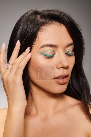 brunette asian woman with green eye makeup and bare shoulders posing on grey background, closed eyes