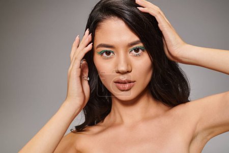 brunette asian woman with sparkling green eye makeup and bare shoulders posing on grey background