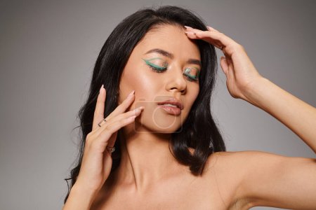 schöne asiatische Frau mit funkelnden grünen Augen Make-up und nackten Schultern posiert auf grauem Hintergrund