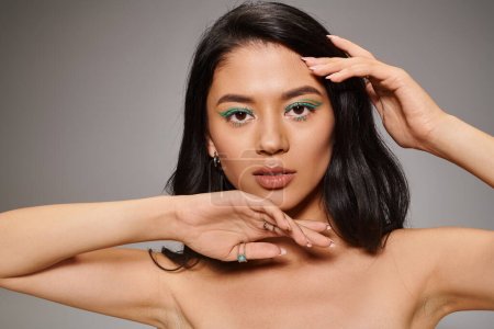 beautiful asian woman with shimmery green eye makeup and bare shoulders posing on grey background