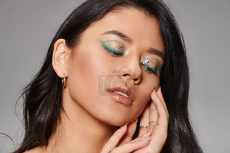 woman with shimmery green makeup and bare shoulders posing with hands near face on grey background