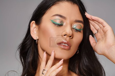 woman with shiny green makeup and bare shoulders posing with hands near face on grey background