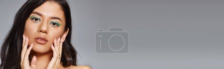 pretty woman with green makeup and bare shoulders posing with hands near face on grey, banner