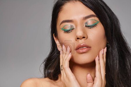 charming woman with green makeup and bare shoulders posing with hands near face on grey background