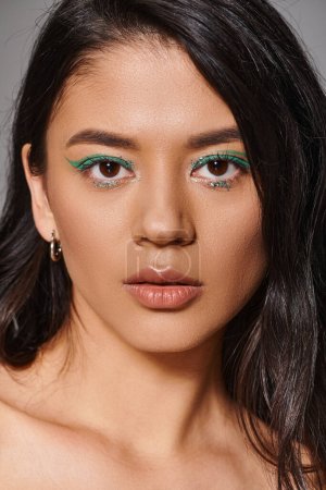 Porträt der schönen Asiatin mit brünetten Haaren und schimmerndem Augen-Make-up auf grauem Hintergrund