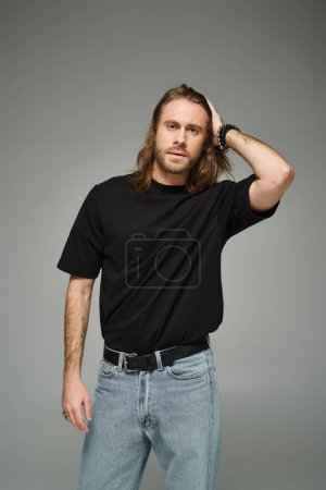 bärtiger, gutaussehender Mann in schwarzem T-Shirt und Jeans mit langen Haaren und grauem Hintergrund