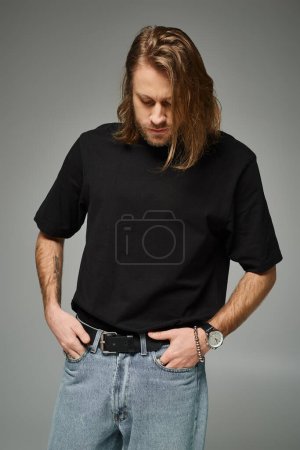 bärtiger und gutaussehender Mann mit langen Haaren posiert in schwarzem T-Shirt und Jeans auf grauem Hintergrund