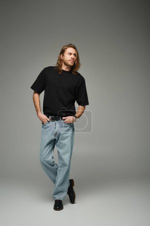 volle Länge des bärtigen gutaussehenden Mannes in schwarzem T-Shirt und Jeans stehend mit der Hand in den Taschen auf grau
