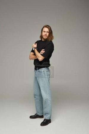 Vollbärtiger und gutaussehender Mann mit langen Haaren posiert in Jeans und T-Shirt vor grauem Hintergrund