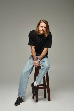 volle Länge des bärtigen und gut aussehenden Mannes mit langen Haaren sitzt in Jeans und T-Shirt auf einem hohen Hocker