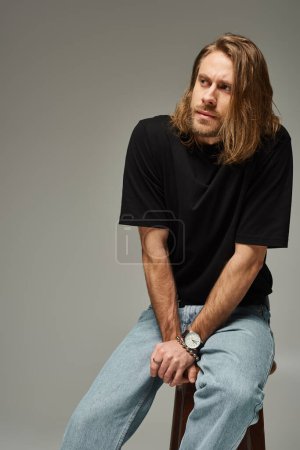 Porträt eines bärtigen und gut aussehenden Mannes mit langen Haaren in Jeans und T-Shirt, der auf einem Stuhl auf grau sitzt