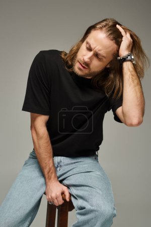 Téléchargez les photos : Portrait d'un homme barbu et beau aux cheveux longs en jean et t-shirt assis sur une chaise grise - en image libre de droit