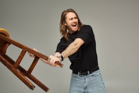 emotional man with long hair in jeans and t-shirt carrying high stool and screaming on grey