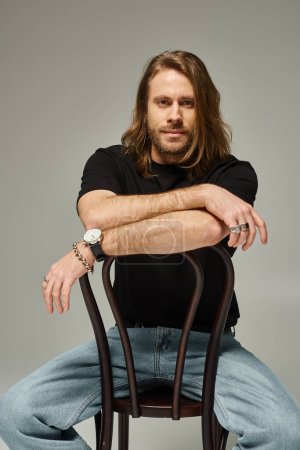 bärtiger, gutaussehender Mann mit langen Haaren sitzt in Jeans und T-Shirt auf einem Holzstuhl in grau