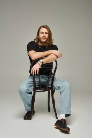 volle Länge des bärtigen gut aussehenden Mannes mit langen Haaren sitzt in Jeans und T-Shirt auf Stuhl auf grau