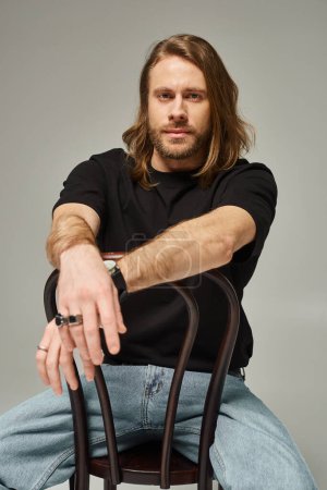 bärtiger und gutaussehender Mann mit langen Haaren sitzt in Jeans und T-Shirt auf Stuhl vor grauem Hintergrund