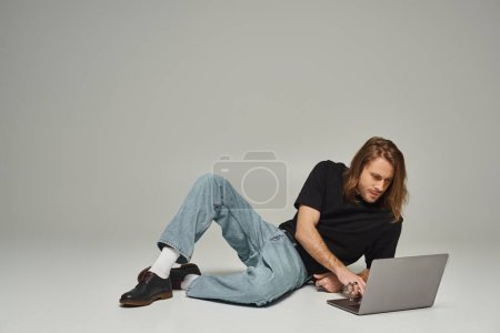 handsome man with long hair and beard sitting in denim jeans on floor and using laptop on grey