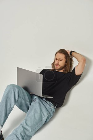 handsome man with long hair and beard leaning on white wall and using laptop on grey backdrop