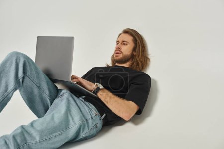handsome man with long hair and beard leaning on white wall and using laptop on grey backdrop
