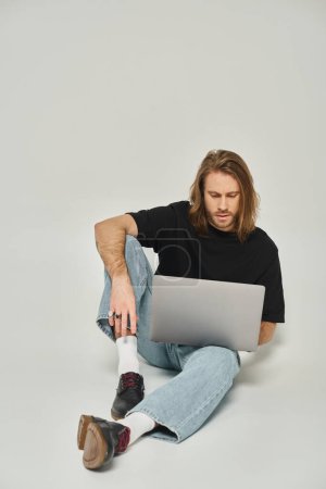 handsome freelancer with long hair and beard sitting on floor and using laptop on grey background