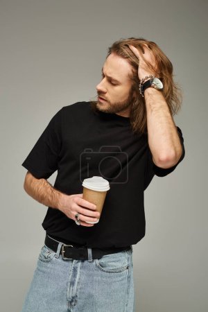 handsome man with long hair and beard holding paper cup with coffee to go on grey background