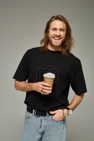 happy man with long hair and beard holding paper cup with coffee to go on grey background