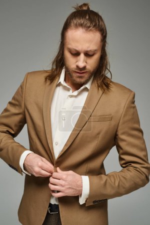 handsome businessman with beard and long hair buttoning blazer on grey backdrop, formal attire