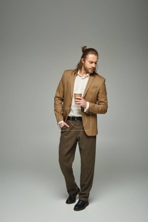 handsome businessman with beard and long hair holding coffee to go on grey backdrop, formal attire
