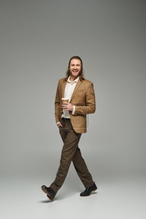 happy businessman with beard and long hair holding coffee to go on grey backdrop, formal attire