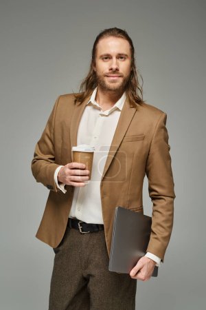 bel homme d'affaires avec barbe et cheveux longs tenant café pour aller et ordinateur portable sur fond gris