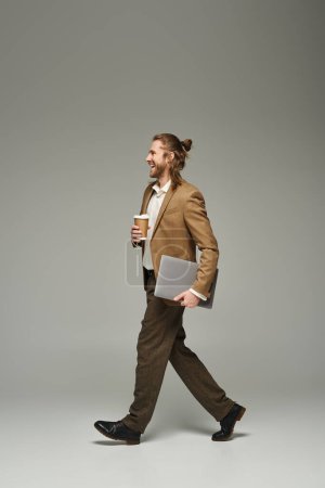feliz hombre de negocios con barba y cabello largo sosteniendo café para llevar y portátil mientras camina sobre gris