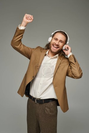 excitado hombre de negocios barbudo en auriculares escuchando música sobre fondo gris, atuendo formal