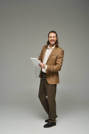 glücklicher Geschäftsmann mit langen Haaren posiert in formeller Kleidung und mit digitalem Tablet vor grauem Hintergrund