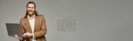 happy businessman with long hair posing in formal attire and using laptop on grey backdrop, banner