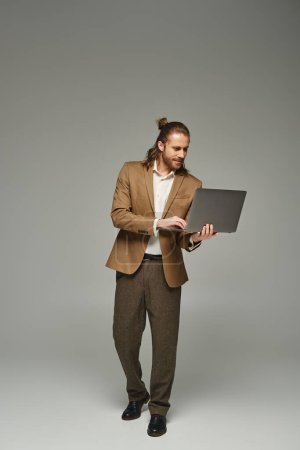 hombre de negocios guapo con el pelo largo posando en traje formal y usando el ordenador portátil en fondo gris