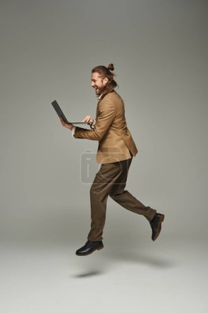 happy businessman with long hair levitating in formal attire and using laptop on grey backdrop