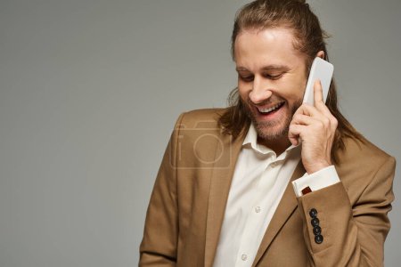 handsome and happy bearded businessman in formal attire talking on smartphone on grey backdrop