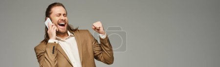 homme d'affaires barbu beau et excité en tenue formelle parlant sur smartphone sur gris, bannière