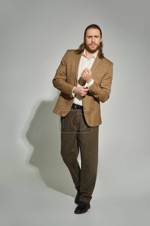 handsome businessman with long hair and beard adjusting sleeve on beige blazer on grey backdrop