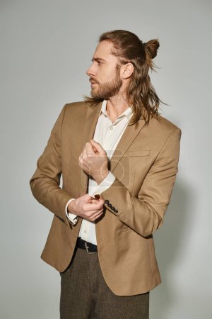 good looking businessman with long hair and beard adjusting sleeve on beige blazer on grey backdrop